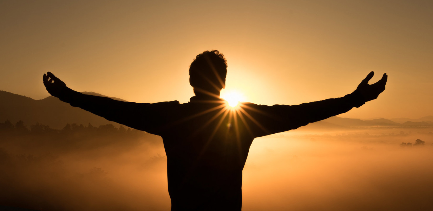 Black silhouette of a man standing on a high peak with arms outstretched overlooking mountains and mist at sunset.