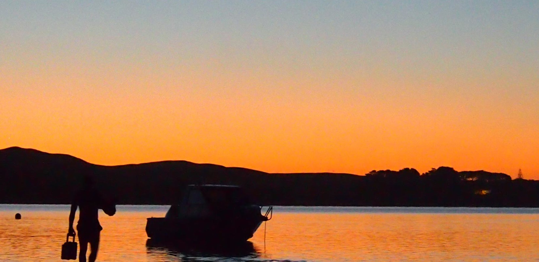 sunset lanscape image with a black silhouette of a man walking to his boat which is moored on the beach.