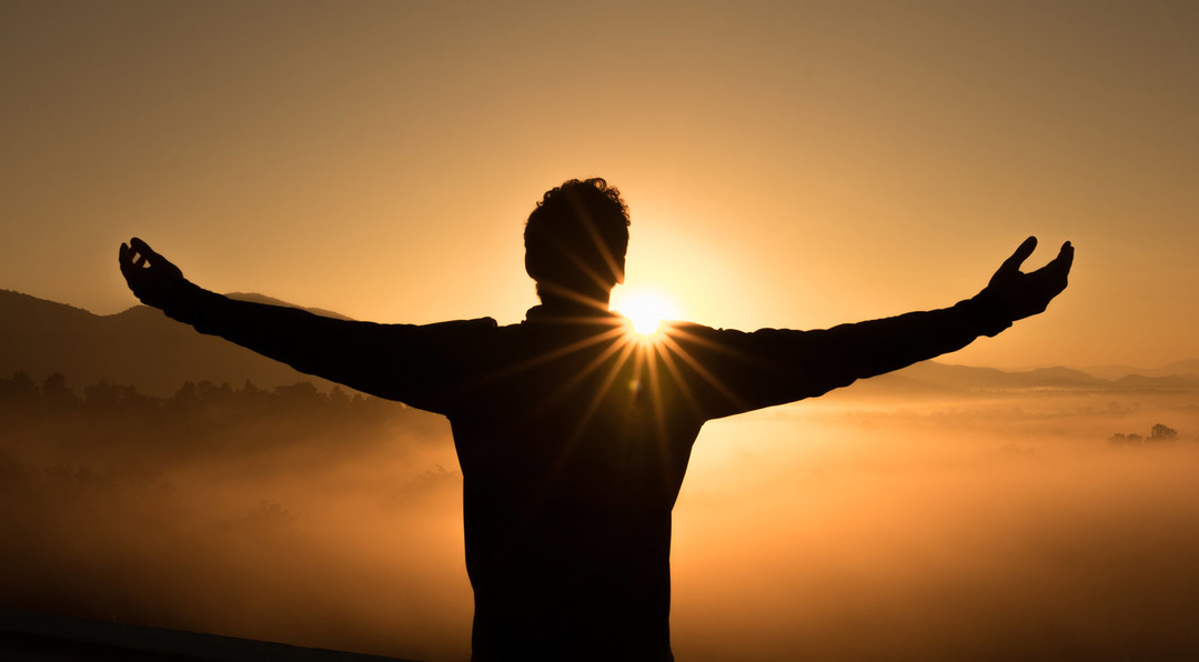 Black silhouette of a man standing on a high peak with arms outstretched overlooking mountains and mist at sunset.